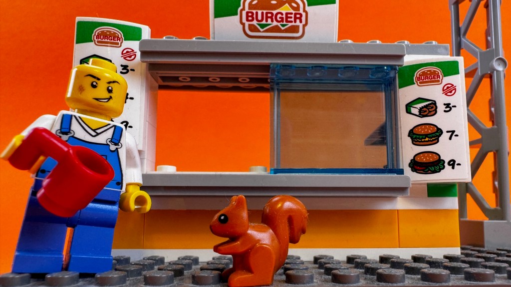 Lego-hahmo punainen kahvikuppi kädessään. Orava katsoo hahmoa. Taustalla hampurilaisgrilli.